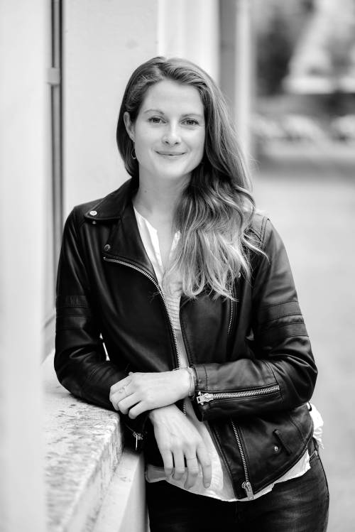 Frau mit langen Haaren in einer Lederjacke, die an einer Leiste lehnt und lächelt, in einer schwarz-weißen Außenumgebung.