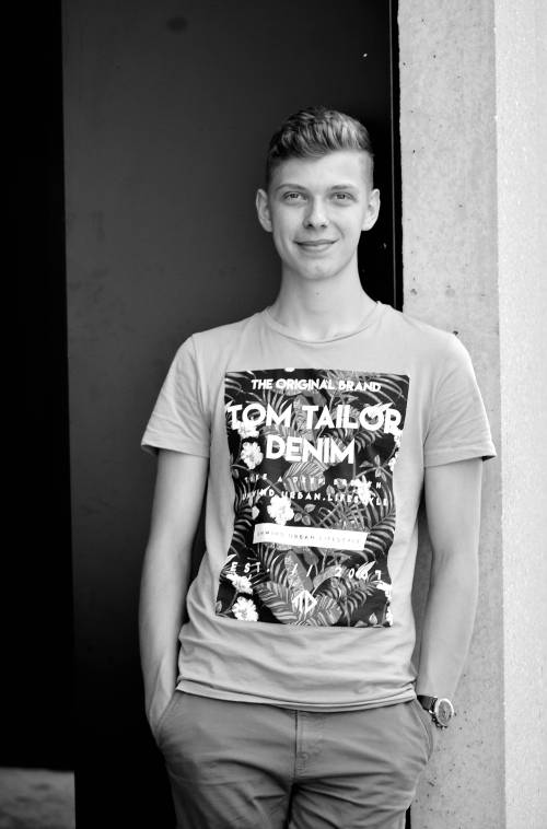 Ein Mann in einem T-Shirt von „Tom Tailor Denim“ lehnt mit den Händen in den Taschen an einer Wand und lächelt auf einem Schwarz-Weiß-Foto in die Kamera.