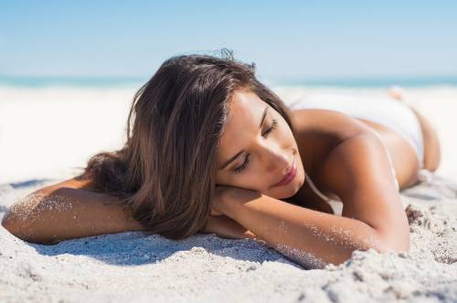 Woman in a white swimsuit lying on sandy beach, resting her head on her arms, with eyes closed, under a clear blue sky.