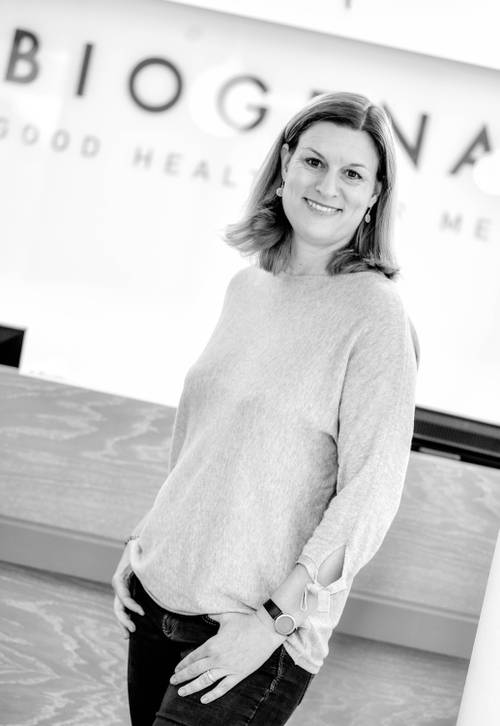 Lächelnde Frau in einem leichten Pullover und Jeans steht lässig vor einer Rezeption, auf der teilweise das Wort „BIOGENA” zu sehen ist.