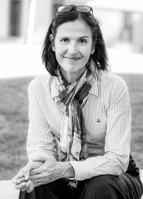 Schwarz-Weiß-Foto einer lächelnden Person mit einer Brille auf dem Kopf, die ein gestreiftes Hemd und einen Schal trägt und im Freien sitzt.