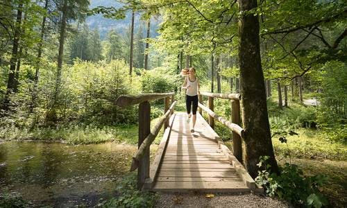 A woman jogs across a wooden bridge in a lush, sun-drenched forest with a small stream flowing beneath it.