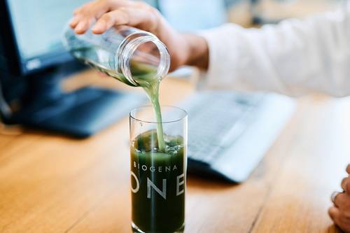 Person, die grünen Saft aus einer Flasche in ein Glas mit Markenlogo auf einem Holztisch einschenkt, im Hintergrund eine Tastatur und ein Monitor