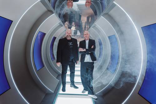 Zwei Männer stehen in einem futuristischen Tunnel mit blauen Paneelen und einer rauchigen Atmosphäre, umgeben von kreisförmigen, beleuchteten Rahmen.