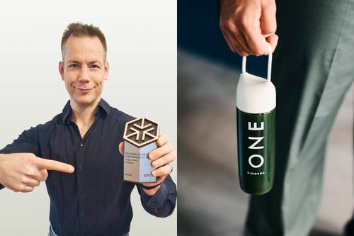 Niekto v modrej košeli ukazuje na šesťuholníkové ocenenie. V druhej ruke drží zelenú fľašu s nápisom „ONE“, ktorá má biely uzáver a rukoväť.