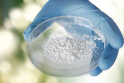 Person holding a petri dish with white powdery substance, wearing a blue glove. Blurred green and white background.