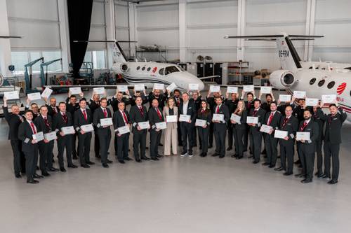 Eine Gruppe von Leuten in formeller Kleidung hält Urkunden in den Händen und steht in einem Hangar, im Hintergrund sind zwei kleine Jets zu sehen.