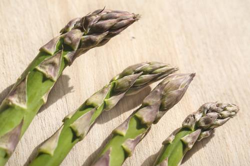 Nahaufnahme von vier frischen Spargelstangen auf einer Holzoberfläche, die ihre grünen Stiele und violetten Knospen zeigen.