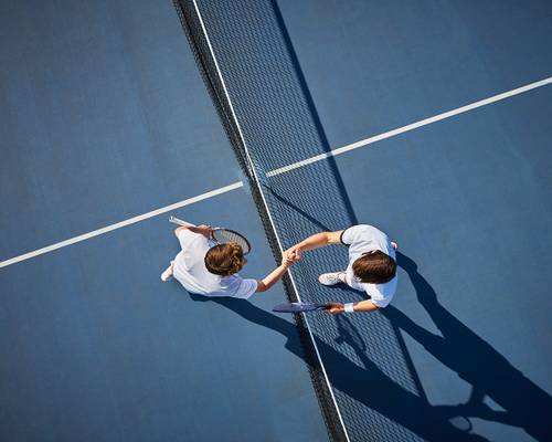 Zwei Tennisspieler in weißen Outfits geben sich über dem Netz auf einem blauen Platz die Hand und werfen dabei lange Schatten.