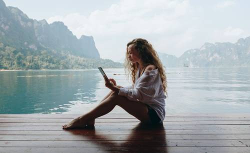 Frau mit langen Haaren sitzt auf einem Holzdeck an einem See und liest auf einem Tablet. Im Hintergrund sind Berge und ein bewölkter Himmel.