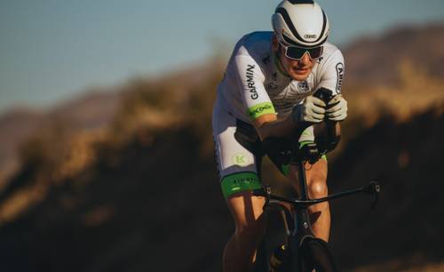 Cyclist in aerodynamic gear riding a road bike through a desert landscape, focused on speed and endurance.