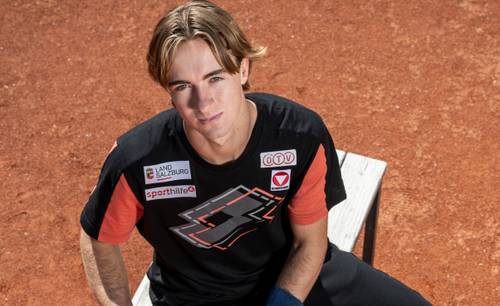 Junger Mann mit hellbraunen Haaren sitzt auf einer Bank auf einem Asche-Sportplatz und trägt ein schwarzes Sportshirt mit Logos und roten Ärmeln.