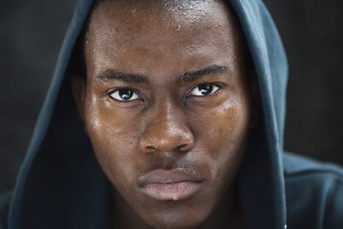 Nahaufnahme einer Person mit Kapuze, ernstem Gesichtsausdruck und Schweißperlen im Gesicht.