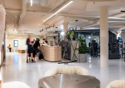 Modernes Fitnessstudio-Interieur mit Personen an der Rezeption, Trainingsgeräten im Hintergrund und Sitzbereich im Vordergrund.