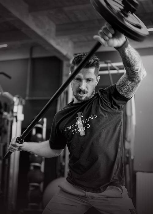 Marcel Hirscher ridică o halteră cu greutăți într-o sală de sport, concentrat pe exercițiul său. Poartă un tricou "The Mountain Studio".