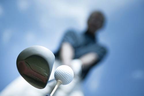 Niedrige Kameraeinstellung eines Golfers, der sich auf den Abschlag vorbereitet, mit Fokus auf den Golfschläger und den Ball vor einem klaren blauen Himmel.