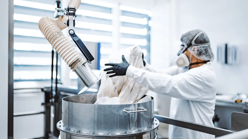Ein Arbeiter in Schutzausrüstung gießt eine Substanz in einen großen Industriekessel in einer hellen, modernen Einrichtung.
