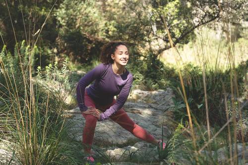 Eine Frau in einem violetten Oberteil und roten Leggings streckt sich im Freien auf einem felsigen Weg, umgeben von Grün und Bäumen.
