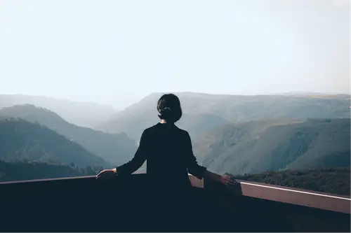 Eine Person in dunkler Kleidung steht auf einem Balkon und blickt unter klarem Himmel auf eine weitläufige Berglandschaft.