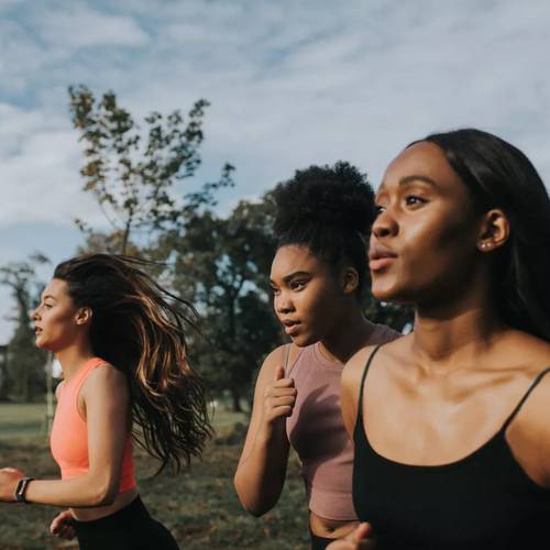 Drei Frauen joggen draußen in Sportkleidung, mit Bäumen und einem bewölkten Himmel im Hintergrund.