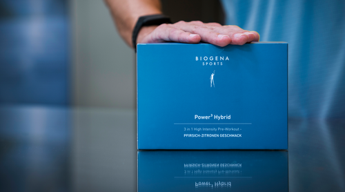 Jemand, der eine Smartwatch trägt, legt seine Hand auf eine blaue Schachtel mit der Aufschrift «BIOGENA SPORTS Power Hybrid», ein Pre-Workout-Supplement mit Zitronengeschmack.