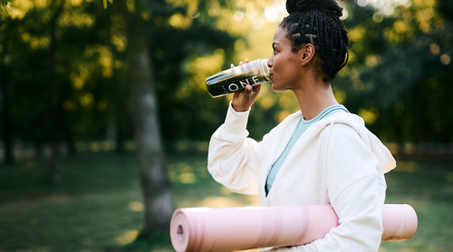 Person trinkt aus BIOGENA ONE Flasche, hält pinke Yogamatte im Park, weißer Hoodie, geflochtene Haare, grüner Hintergrund.