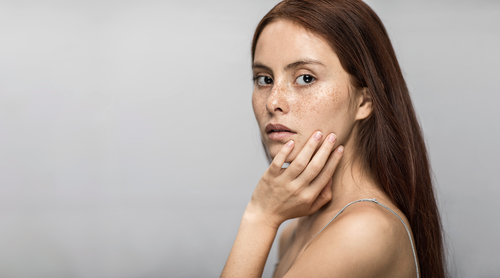 Woman with long red hair and freckles, touching her face, gazes thoughtfully at the camera against a plain gray background.