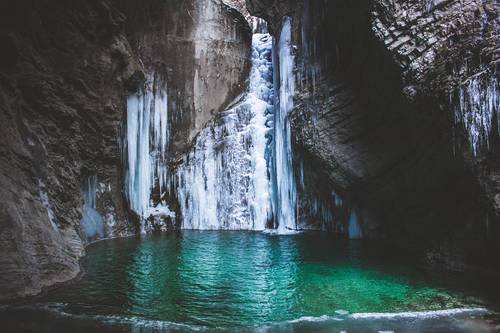Ein gefrorener Wasserfall, der in ein türkisfarbenes Becken stürzt, umgeben von eisigen und felsigen Klippen.