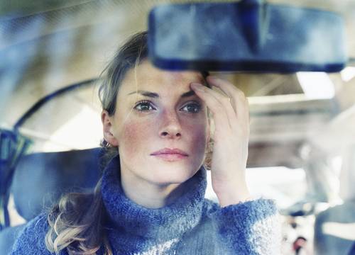 Eine Frau in einem Auto justiert den Rückspiegel, trägt einen blauen Pullover und blickt konzentriert und nachdenklich durch die Windschutzscheibe.