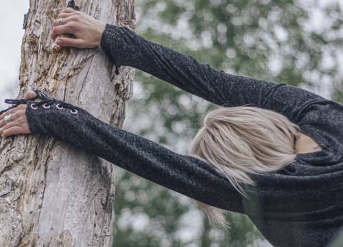 Person with blonde hair stretching against a tree, wearing a dark long-sleeve top, with a blurred green background.