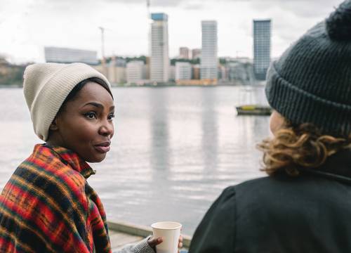 Zwei Personen in Winterkleidung unterhalten sich am Ufer, im Hintergrund sind Stadtgebäude zu sehen. Eine Person hält eine Tasse in einer karierten Decke.