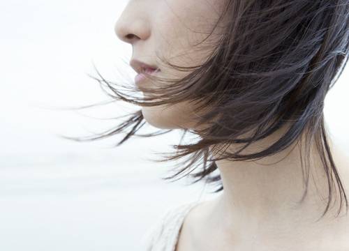 Nahaufnahme des Gesichts einer Person im Profil mit kurzen, dünnen Haaren, die im Wind wehen, vor einem weichen, unscharfen Hintergrund.