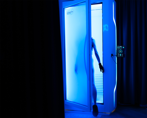 Person, die eine blau beleuchtete Kryotherapiekammer betritt, Handschuhe und Schutzschuhe trägt, mit einem Bedienfeld an der Seite.