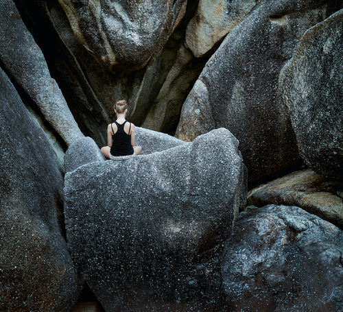 A person in a black tank top sits on large boulders, surrounded by rugged rock formations, with their back facing the camera.