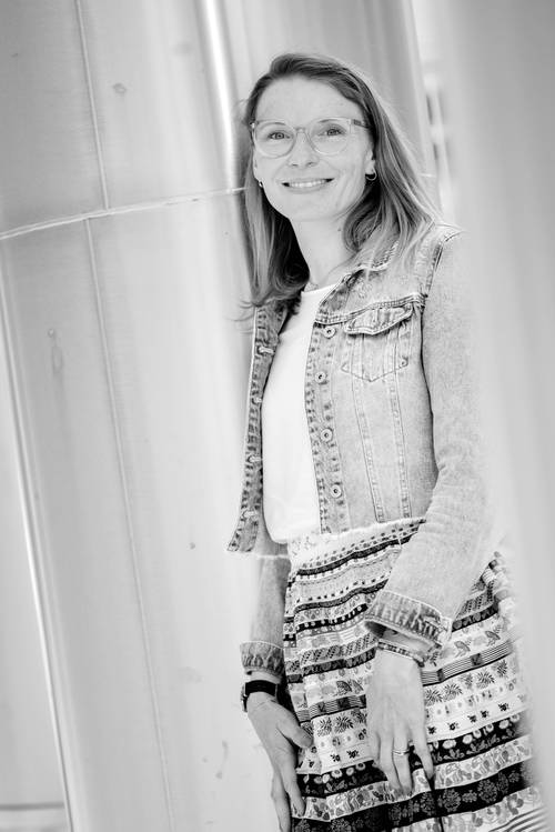 Frau mit Brille und Jeansjacke steht lächelnd da. Trägt einen gemusterten Rock. Schwarz-Weiß-Foto mit Metallpfeiler im Hintergrund.