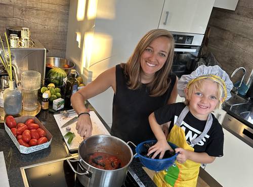 Eine Frau und ein Kind in einer gelben Schürze kochen gemeinsam in einer Küche, lächeln dabei und auf der Arbeitsplatte liegen Tomaten und andere Zutaten.