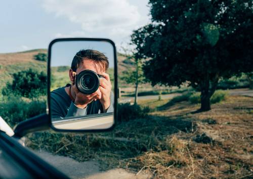 Persoană care face o fotografie prin oglinda laterală a unei mașini, cu un peisaj rural și copaci în fundal.