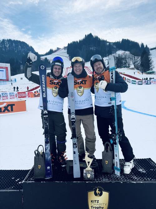 Three skiers in helmets and uniforms pose on a podium with skis and a trophy, in a snowy mountain setting with event banners behind them.