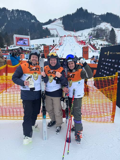 Three skiers in orange vests pose with their ski gear at a snowy slope, with a cheering crowd and mountainous backdrop.