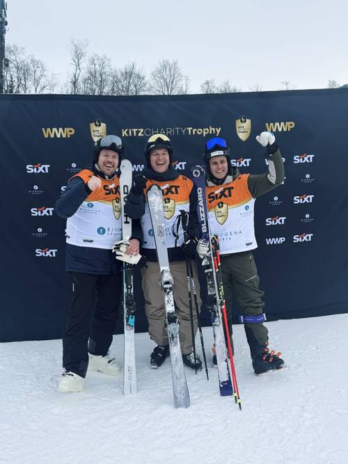 Three skiers in matching outfits celebrate in front of a KitzCharity Trophy backdrop, holding skis and smiling on a snowy slope.