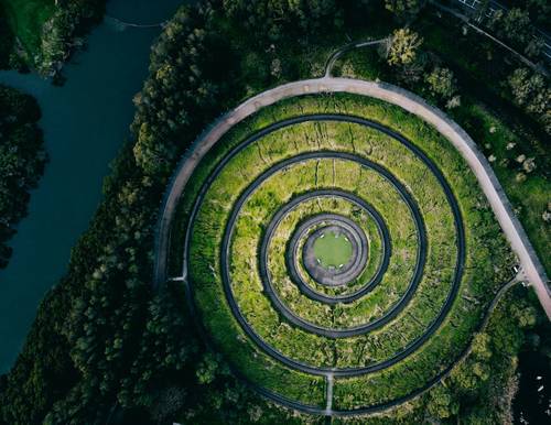 Vedere aeriană a unei grădini circulare în terase, cu alei concentrice, înconjurată de vegetație deasă și un râu din apropiere.