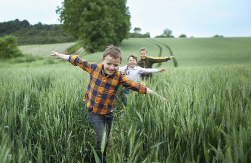Drei Kinder laufen fröhlich über eine üppig grüne Wiese, wobei eines der Kinder im Vordergrund ein kariertes Hemd trägt. Im Hintergrund sind ein Baum und Hügel zu sehen.