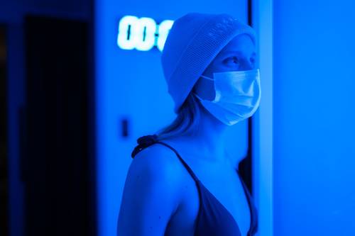 Person in einer Maske und Mütze, erleuchtet von blauem Licht, steht in einem schwach beleuchteten Raum in der Nähe eines Digitaluhr, die "00:12" anzeigt.