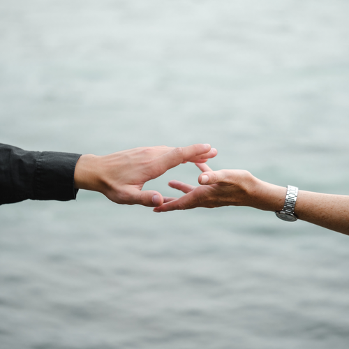 Zwei Hände, die sich vor einem verschwommenen Wasserhintergrund entgegenstrecken. An einer Hand steckt eine Uhr, die andere steckt in einem dunklen Ärmel.