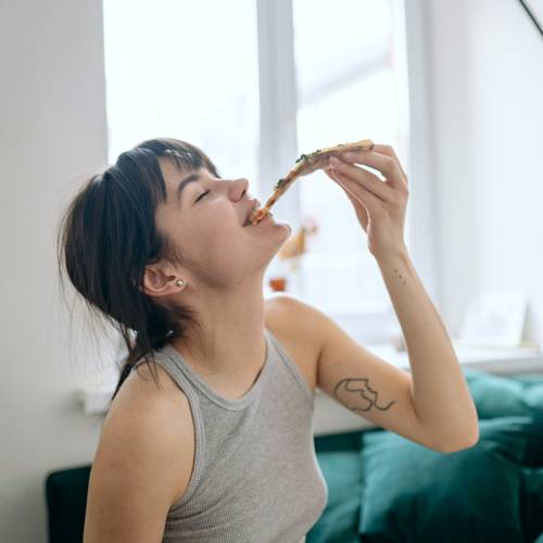 Eine Frau im grauen Tanktop isst drinnen fröhlich ein Stück Pizza, mit einem Fenster im Hintergrund.