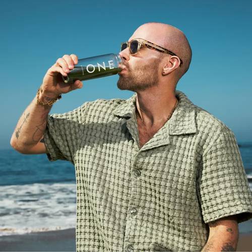 Ein Mann in Sonnenbrille trinkt aus einer Flasche mit dem Etikett "ONE" am Strand, während er ein strukturiertes grünes Hemd trägt. Der Ozean und der Himmel sind im Hintergrund.