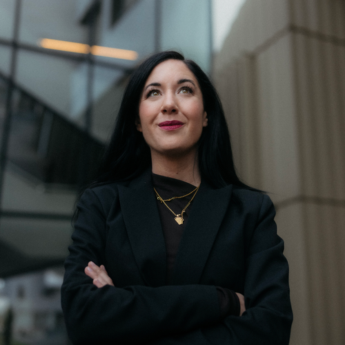 Julia Hoffmann într-un blazer negru cu brațele încrucișate, stând în fața unei clădiri moderne, privind în sus cu o expresie încrezătoare.