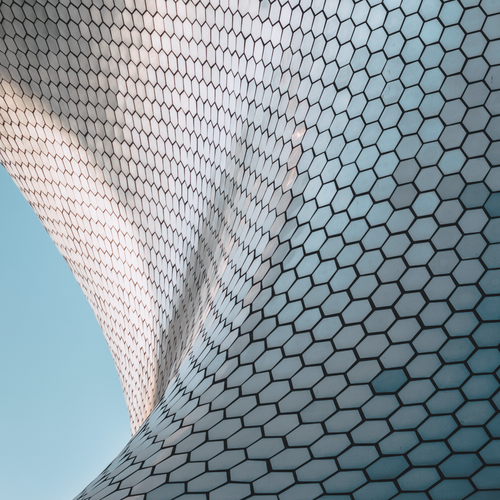 O fațadă curbată a clădirii, acoperită cu plăci hexagonale, care reflectă lumina pe fundalul unui cer albastru strălucitor, creând astfel un model modern și abstract.