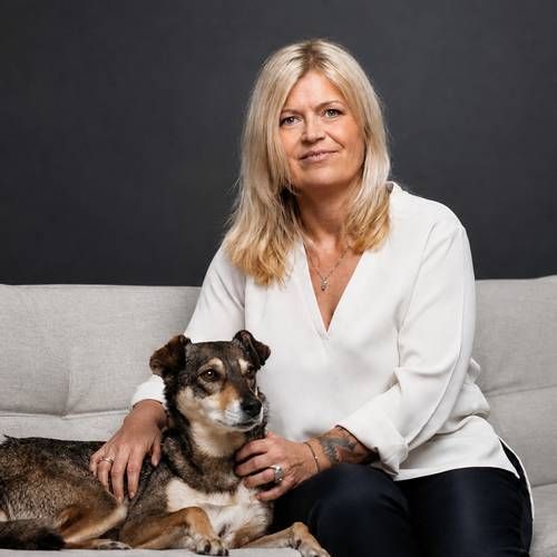 BIOGENA PETS Tierärztin sitzt auf einem grauen Sofa und hält sanft einen Hund. Beide blicken in die Kamera vor einem dunklen Hintergrund.
