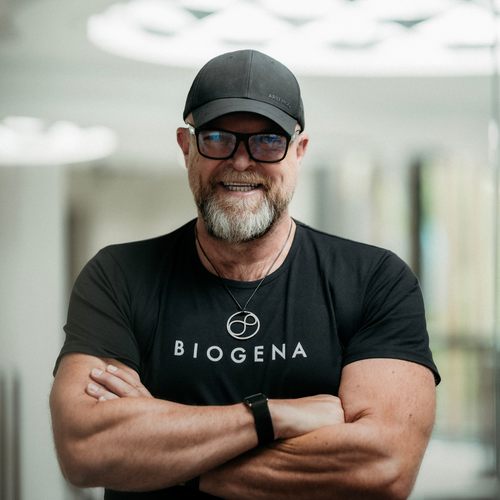 Albert Schmidbauer cu barbă, șapcă neagră și ochelari, zâmbește cu brațele încrucișate, purtând un tricou negru cu logo-ul "BIOGENA".